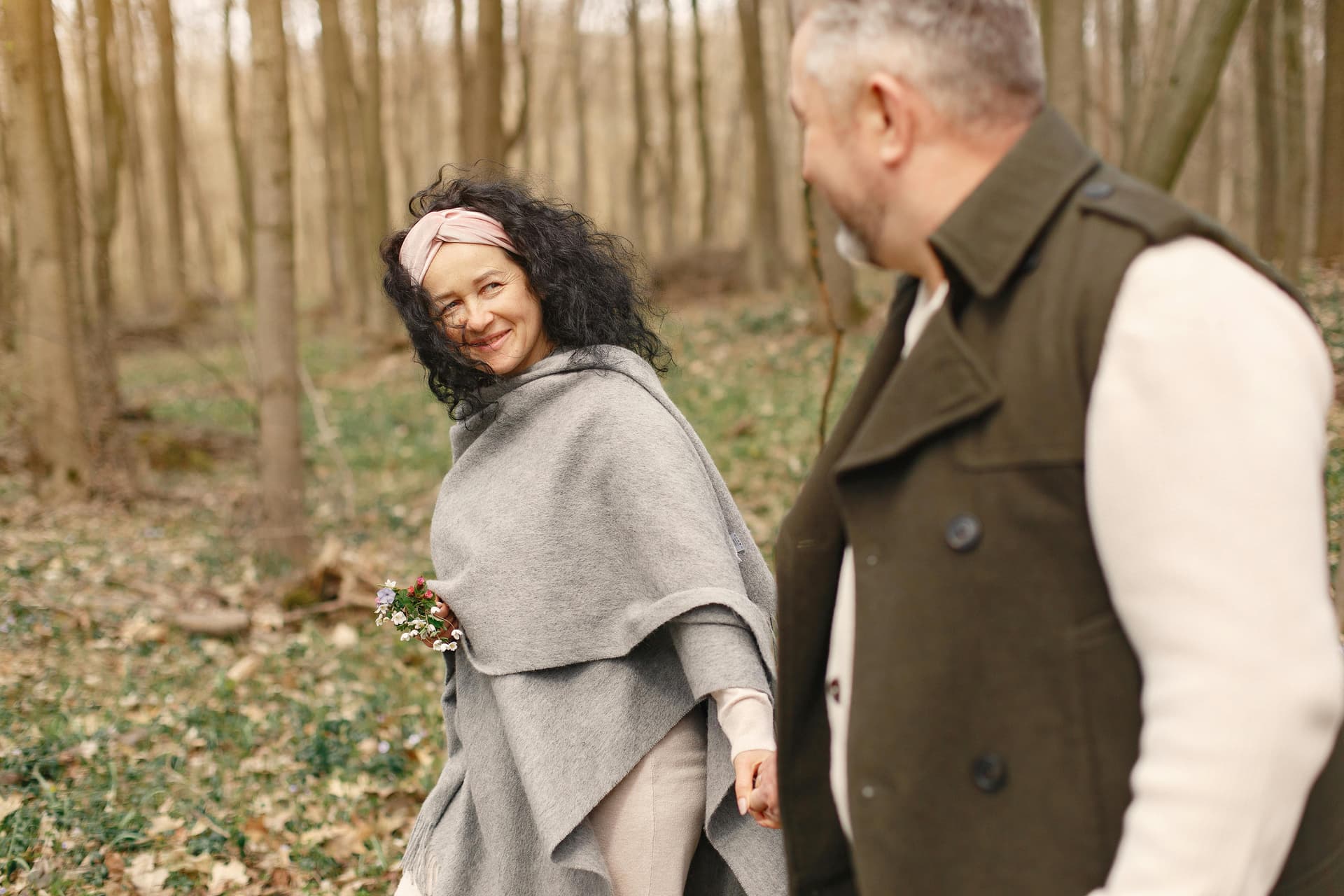 A woman in a gray poncho and a headband smiles at a man while walking in a forest.