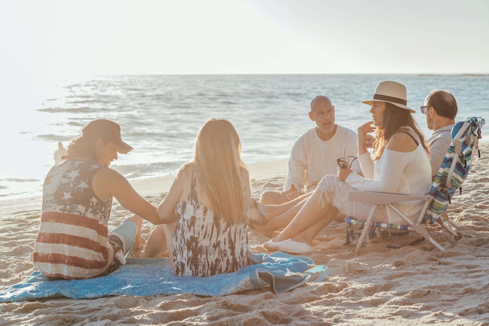 A group of five people sits on the beach, engaged in conversation with the ocean in the background.