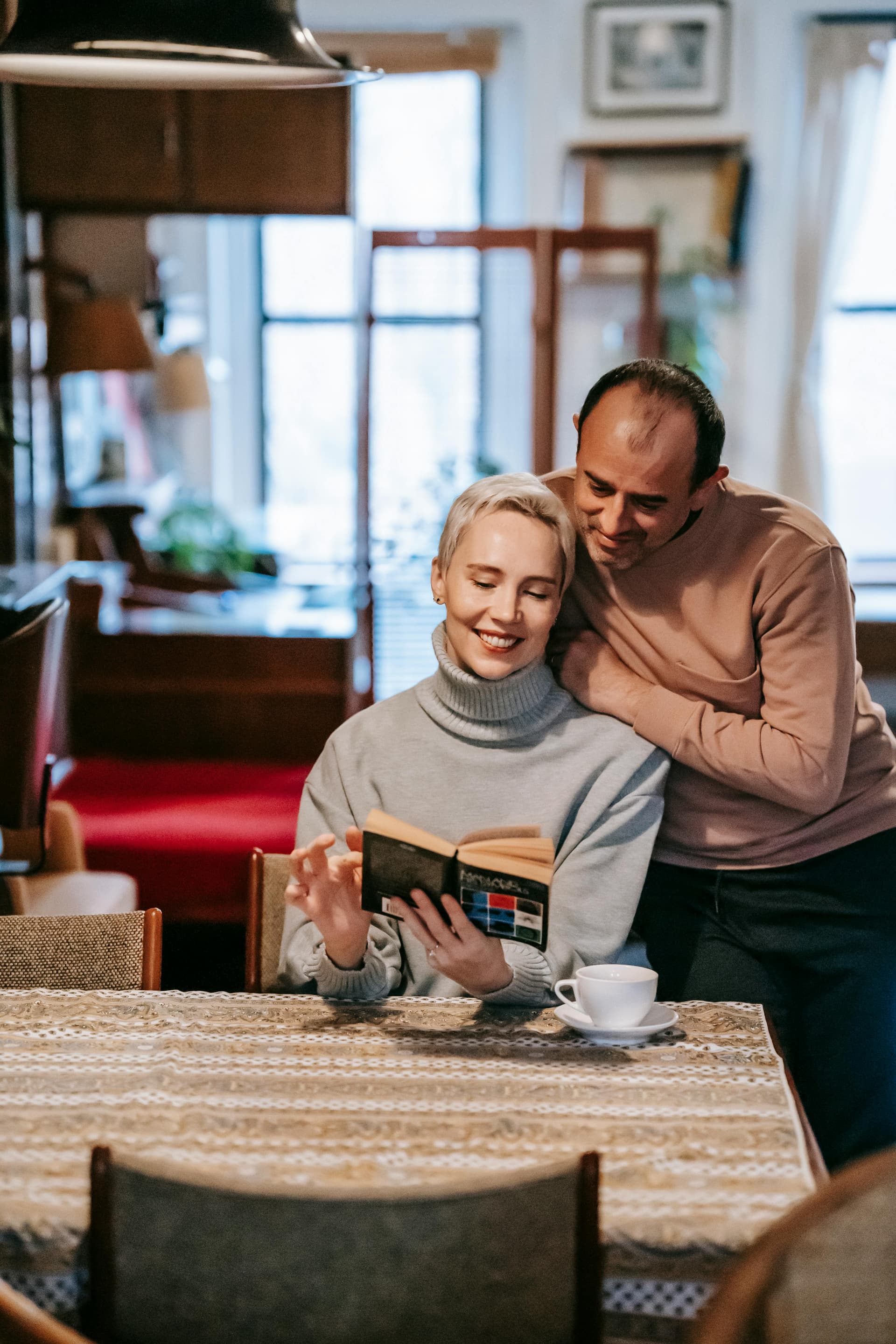 A woman reads a book while a man embraces her from behind in a cozy indoor setting.